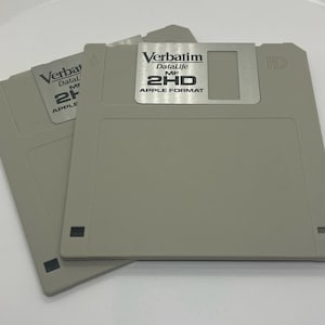 May include: Two gray Verbatim DataLife 2HD Apple Format floppy disks. The disks have a rectangular shape with a metal label and black square details. The text "Verbatim DataLife MF 2HD APPLE FORMAT" is visible on the label.