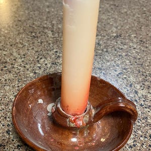 May include: A brown ceramic candle holder with a handle, holding a tall, partially burned, peach-colored candle. The candle has red wax drippings. The candle holder has a round base and a small, circular holder for the candle.