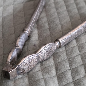 May include: Silver nutcracker with ornate floral and scrollwork designs. The handles and body of the nutcracker are detailed with embossed patterns. The nutcracker is open, ready for use, and sits on a textured surface.