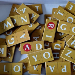 May include: A collection of Scrabble tiles, featuring yellow tiles with white letters and numbers. Some tiles have red circles with white letters. The tiles are scattered, showing various letters and point values.