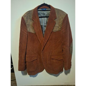 May include: A brown corduroy blazer with suede shoulder patches and elbow patches. The blazer has two front pockets and is hanging on a black hanger. The interior lining is a gray color.