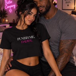 May include: A black short-sleeved crop top featuring the white text PUNISH ME, and the pink text DADDY. The garment has a crew neckline and a cropped hemline, constructed from a solid black fabric with a simple, fitted silhouette.