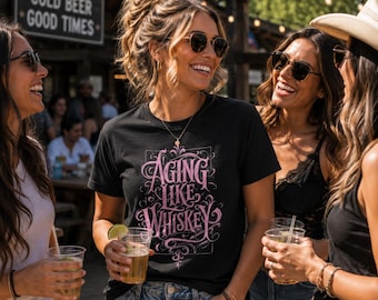May include: A black crew neck t-shirt featuring pink decorative script text that reads Aging Like Whiskey, centered on the chest. The shirt is made of a solid black fabric with a screen-printed graphic design in a light pink color.