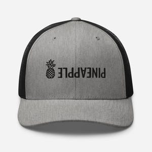 Puede incluir: Una gorra de camionero gris y negra con un gráfico de piña negro y la palabra "PINEAPPLE" escrita al revés.