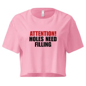 Könnte beinhalten: Ein hellrosa bauchfreies T-Shirt mit Rundhalsausschnitt und kurzen Ärmeln. In der Mitte des T-Shirts befindet sich der Text ATTENTION! in Rot, gefolgt von den Worten HOLES NEED FILLING in fetten schwarzen Großbuchstaben auf der Vorderseite.