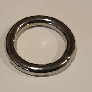 Può includere: Un anello di metallo argentato lucido. L'anello è circolare con una superficie liscia e riflettente. L'anello è un pezzo di metallo solido e senza cuciture.