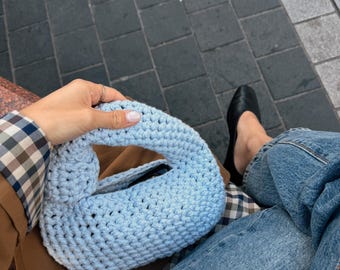 Puede incluir: Un bolso de ganchillo azul claro con un diseño de asa anudada. El bolso está confeccionado con un hilo grueso y texturizado en un patrón de punto sencillo, creando una forma redondeada y compacta con una abertura superior.