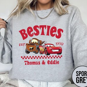 May include: Sport grey sweatshirt featuring a graphic of two cartoon cars with the text "Besties" and "Thomas & Eddie". The design includes the year "1996" and a checkered pattern. The sweatshirt is a crew neck.