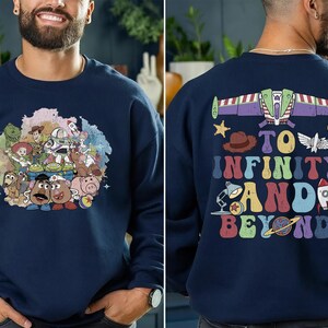 May include: Navy blue sweatshirt featuring a colorful graphic of Toy Story characters on the front. The back of the sweatshirt displays a Buzz Lightyear design with the phrase "To Infinity and Beyond" in a retro font.