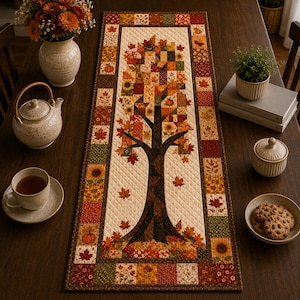 May include: A rectangular quilted table runner featuring a central tree motif with autumn-colored leaves, surrounded by a border of patchwork squares in shades of orange, brown, yellow, and green, with a cream-colored background and diamond stitching.