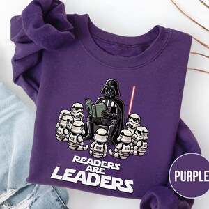 K&ouml;nnte beinhalten: Ein lila Sweatshirt mit einer Grafik von Darth Vader, der Stormtroopern vorliest. Der Text "READERS ARE LEADERS" ist unter der Illustration aufgedruckt. Ein lila Kreis mit dem Wort "PURPLE" befindet sich in der unteren rechten Ecke.