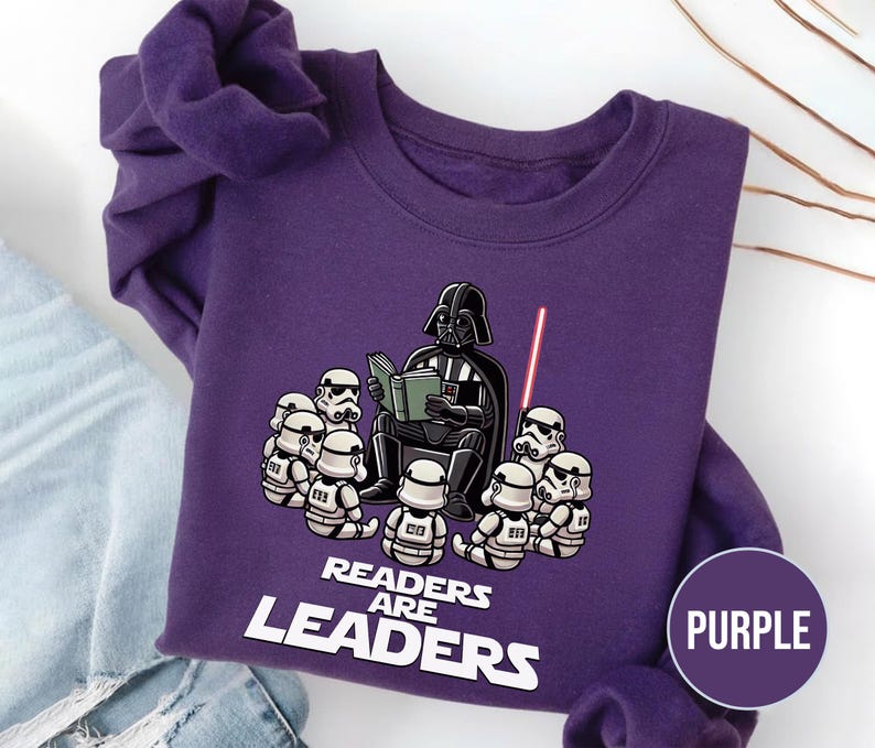 K&ouml;nnte beinhalten: Ein lila Sweatshirt mit einer Grafik von Darth Vader, der Stormtroopern vorliest. Der Text "READERS ARE LEADERS" ist unter der Illustration aufgedruckt. Ein lila Kreis mit dem Wort "PURPLE" befindet sich in der unteren rechten Ecke.