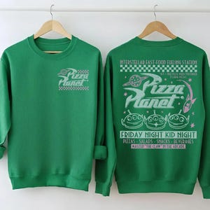 May include: Green sweatshirt featuring the Pizza Planet logo on the front and back. The back design includes the text "Interstellar Fast-Food Fueling Station" and "Friday Night Kid Night". The design also features three green alien characters.
