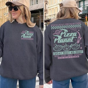 May include: Dark gray Pizza Planet sweatshirt with a front and back graphic. The front graphic features the Pizza Planet logo with a checkered border. The back graphic includes the Pizza Planet logo, aliens, and text about the restaurant.