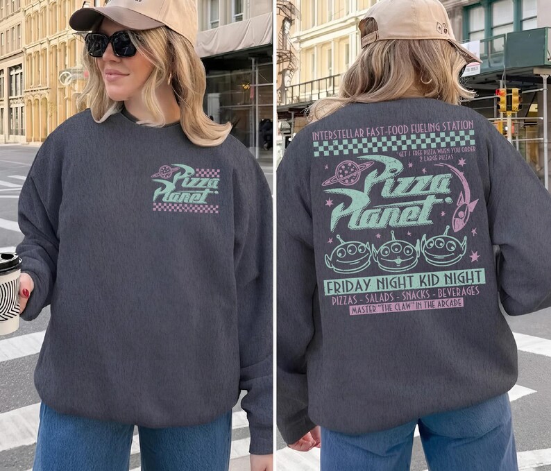 May include: Dark gray Pizza Planet sweatshirt with a front and back graphic. The front graphic features the Pizza Planet logo with a checkered border. The back graphic includes the Pizza Planet logo, aliens, and text about the restaurant.