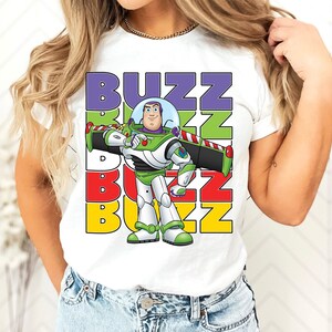 Puede incluir: Camiseta blanca con un gr&aacute;fico de Buzz Lightyear de Toy Story. El dise&ntilde;o incluye la palabra "BUZZ" en letras apiladas y coloridas. Buzz se representa con su traje espacial verde y blanco.