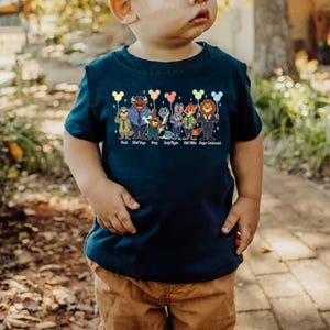 May include: Navy blue toddler t-shirt featuring a colorful graphic of cartoon animal characters holding balloons. The characters include a sloth, a bull, a sheep, a bunny, a fox, and a lion. The names of the characters are printed below them.