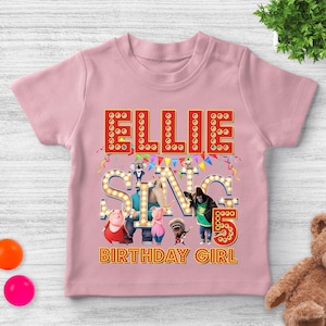 May include: A light pink t-shirt featuring the word "ELLIE" in red marquee letters. Below, the word "SING" is spelled out with light-up letters, and the number "5" is displayed. The shirt also includes the text "BIRTHDAY GIRL" and cartoon characters.