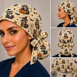 May include: A beige scrub cap featuring a repeating pattern of raccoons, orange pumpkins, black bats, spider webs, and stars. The cap has a gathered back section with a fabric tie closure, designed to secure hair in a ponytail or bun style.