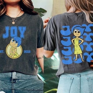 May include: Dark gray t-shirt featuring the word "JOY" in blue block letters with a cartoon character illustration. The back of the shirt has the word "JOY" repeated with the same character. The shirt has a vintage, worn look.