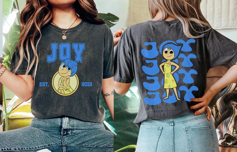 May include: Dark gray t-shirt featuring the word "JOY" in blue block letters with a cartoon character illustration. The back of the shirt has the word "JOY" repeated with the same character. The shirt has a vintage, worn look.