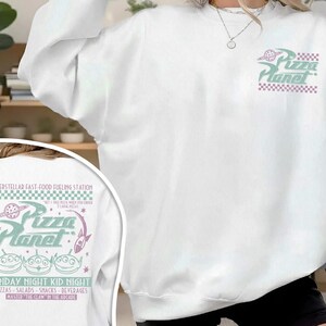 May include: White sweatshirt with a Pizza Planet graphic on the front and back. The front graphic features the Pizza Planet logo in green and purple. The back graphic includes the text "INTERSTELLAR FAST-FOOD FUELING STATION" and "FRIDAY NIGHT KID NIGHT".