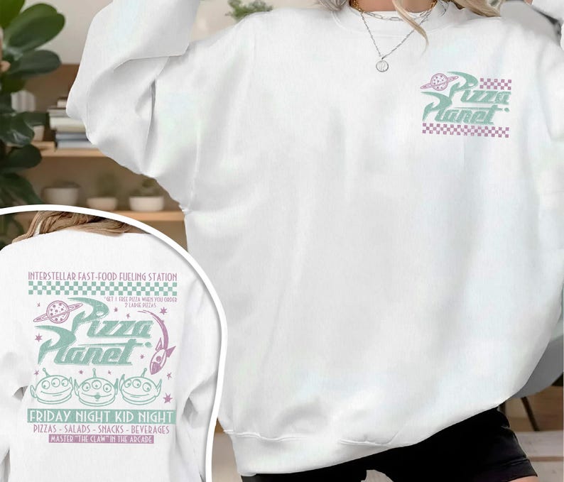 May include: White sweatshirt with a Pizza Planet graphic on the front and back. The front graphic features the Pizza Planet logo in green and purple. The back graphic includes the text "INTERSTELLAR FAST-FOOD FUELING STATION" and "FRIDAY NIGHT KID NIGHT".