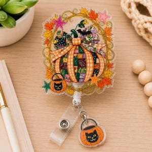 May include: A clear acrylic badge reel featuring a central pumpkin with a patchwork pattern of orange, green, and purple, surrounded by a gold-toned frame of autumn leaves, stars, and a small black cat trick-or-treat bucket charm.