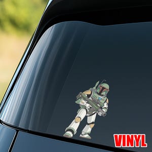 Puede incluir: Una calcoman&iacute;a de vinilo con una ilustraci&oacute;n de dibujos animados de un cazarrecompensas con armadura verde, blanca y gris, sosteniendo un rifle bl&aacute;ster. La palabra "VINYL" est&aacute; impresa en rojo.