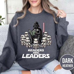 K&ouml;nnte beinhalten: Dunkelgraues Sweatshirt mit einer Grafik von Darth Vader, der Stormtroopern vorliest. Der Text "Readers Are Leaders" ist unter der Illustration aufgedruckt. Ein rotes Lichtschwert ist ebenfalls im Bild.