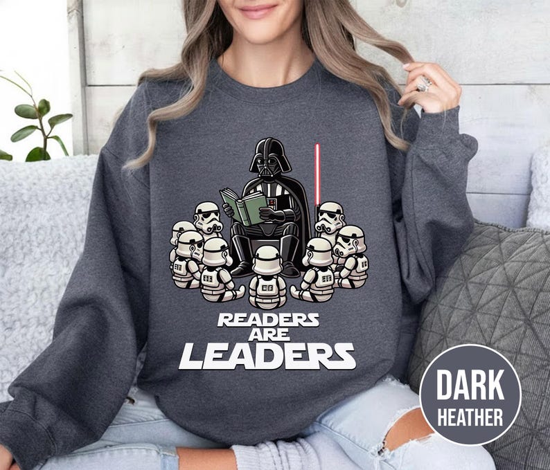 K&ouml;nnte beinhalten: Dunkelgraues Sweatshirt mit einer Grafik von Darth Vader, der Stormtroopern vorliest. Der Text "Readers Are Leaders" ist unter der Illustration aufgedruckt. Ein rotes Lichtschwert ist ebenfalls im Bild.