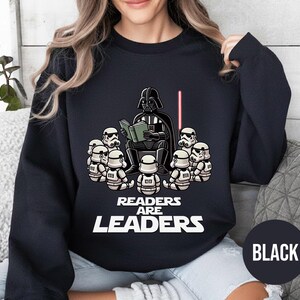 K&ouml;nnte beinhalten: Schwarzes Sweatshirt mit einer Cartoon-Illustration von Darth Vader, der einer Gruppe von Sturmtruppen vorliest. Der Text "READERS ARE LEADERS" ist unter der Illustration aufgedruckt. Ein rotes Lichtschwert ist ebenfalls zu sehen. Das Wort "BLACK" befindet sich in einem schwarzen Kreis.
