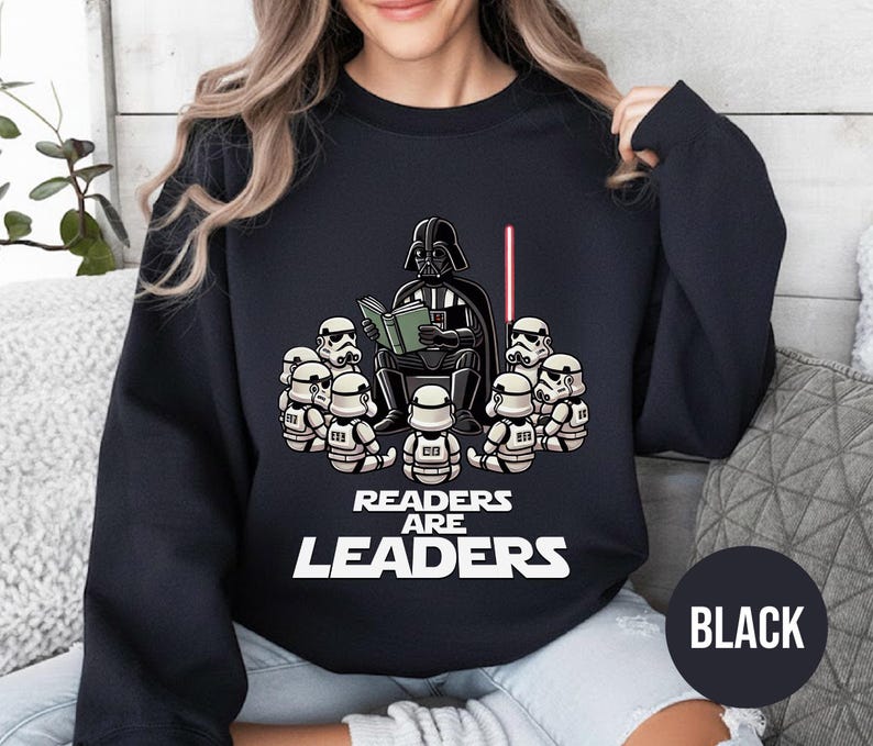 K&ouml;nnte beinhalten: Schwarzes Sweatshirt mit einer Cartoon-Illustration von Darth Vader, der einer Gruppe von Sturmtruppen vorliest. Der Text "READERS ARE LEADERS" ist unter der Illustration aufgedruckt. Ein rotes Lichtschwert ist ebenfalls zu sehen. Das Wort "BLACK" befindet sich in einem schwarzen Kreis.