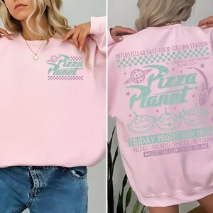 May include: A light pink sweatshirt with the Pizza Planet logo on the front and back. The front features the logo in a teal and purple color scheme. The back has the text "INTERSTELLAR FAST-FOOD FUELING STATION" and "FRIDAY NIGHT KID NIGHT".