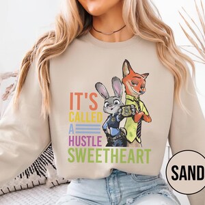 Puede incluir: Sudadera color arena con un gr&aacute;fico colorido de Judy Hopps y Nick Wilde de Zootopia, con el texto "IT'S CALLED A HUSTLE SWEETHEART". El dise&ntilde;o incluye una placa y los personajes con su atuendo de polic&iacute;a y zorro.