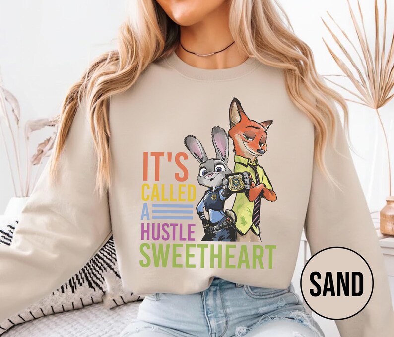 Puede incluir: Sudadera color arena con un gr&aacute;fico colorido de Judy Hopps y Nick Wilde de Zootopia, con el texto "IT'S CALLED A HUSTLE SWEETHEART". El dise&ntilde;o incluye una placa y los personajes con su atuendo de polic&iacute;a y zorro.