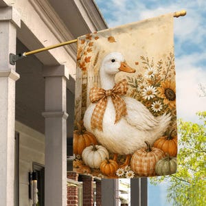 May include: A decorative garden flag featuring a white duck wearing a tan and white checkered bow around its neck. The duck is surrounded by orange and white pumpkins, sunflowers, and daisies against a light beige background with autumn leaves.
