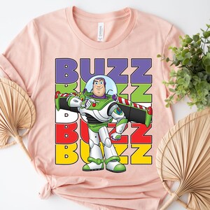 Puede incluir: Una camiseta rosa claro con un dibujo animado de Buzz Lightyear de Toy Story. La palabra "BUZZ" est&aacute; apilada verticalmente en letras coloridas. Buzz Lightyear se representa con su traje espacial verde, blanco y morado.