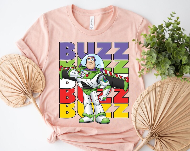 Puede incluir: Una camiseta rosa claro con un dibujo animado de Buzz Lightyear de Toy Story. La palabra "BUZZ" est&aacute; apilada verticalmente en letras coloridas. Buzz Lightyear se representa con su traje espacial verde, blanco y morado.