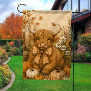 May include: A rectangular garden flag featuring a fluffy brown Highland cow with long horns and a large tan plaid bow around its neck. The cow is surrounded by orange and white pumpkins, white daisies, and falling autumn leaves near a wooden fence.