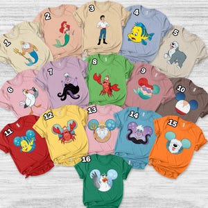 May include: A collection of short-sleeved t-shirts in various colors, each featuring a cartoon character design. Designs include a mermaid, a crab, and a dog. The shirts are arranged on a light wood surface.