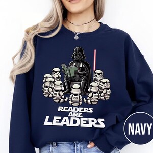 K&ouml;nnte beinhalten: Marineblaues Sweatshirt mit einer Cartoon-Illustration von Darth Vader, der einer Gruppe von Sturmtruppen vorliest. Der Text "READERS ARE LEADERS" ist unter der Illustration aufgedruckt. Ein rotes Lichtschwert ist ebenfalls zu sehen.