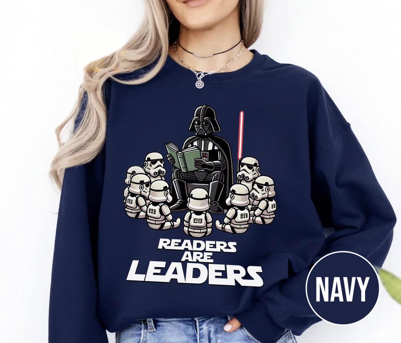 K&ouml;nnte beinhalten: Marineblaues Sweatshirt mit einer Cartoon-Illustration von Darth Vader, der einer Gruppe von Sturmtruppen vorliest. Der Text "READERS ARE LEADERS" ist unter der Illustration aufgedruckt. Ein rotes Lichtschwert ist ebenfalls zu sehen.