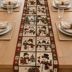 May include: A rectangular fabric table runner featuring a grid of illustrated snowmen in various winter scenes. The design includes text reading JOY and HAPPY HOLIDAYS, surrounded by a red and green border with a repeating snowflake pattern.