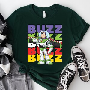 Puede incluir: Camiseta verde oscuro con un gr&aacute;fico de Buzz Lightyear y la palabra "BUZZ" en letras apiladas y coloridas. El dise&ntilde;o muestra al personaje con su traje espacial verde, blanco y morado. Zapatillas de lona negras con cordones blancos junto a la camiseta.