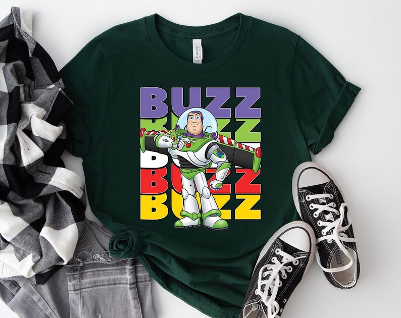 Puede incluir: Camiseta verde oscuro con un gr&aacute;fico de Buzz Lightyear y la palabra "BUZZ" en letras apiladas y coloridas. El dise&ntilde;o muestra al personaje con su traje espacial verde, blanco y morado. Zapatillas de lona negras con cordones blancos junto a la camiseta.
