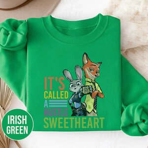 Puede incluir: Sudadera verde irland&eacute;s con un dise&ntilde;o de un zorro y un conejo de dibujos animados y el texto "IT'S CALLED A HUSTLE SWEETHEART". La parte superior de manga larga y cuello redondo tiene una etiqueta circular "IRISH GREEN".