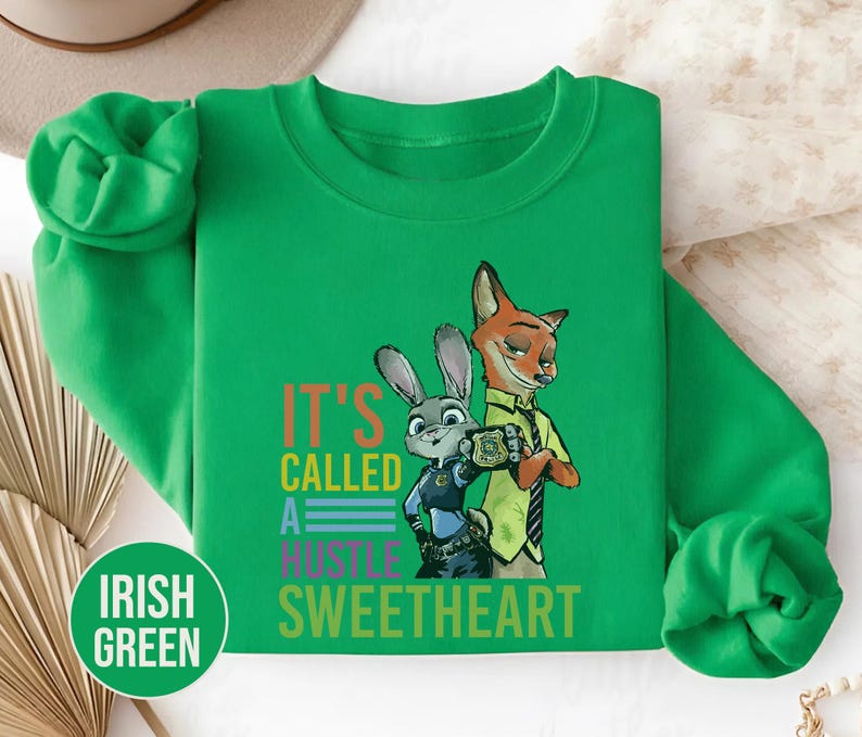 Puede incluir: Sudadera verde irland&eacute;s con un dise&ntilde;o de un zorro y un conejo de dibujos animados y el texto "IT'S CALLED A HUSTLE SWEETHEART". La parte superior de manga larga y cuello redondo tiene una etiqueta circular "IRISH GREEN".