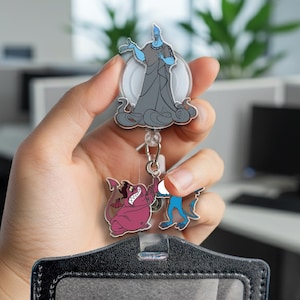 May include: A retractable badge reel featuring cartoon characters. The top character is a blue figure with grey robes. Below are two smaller cartoon figures, one pink and one blue. The reel is attached to a black badge holder.