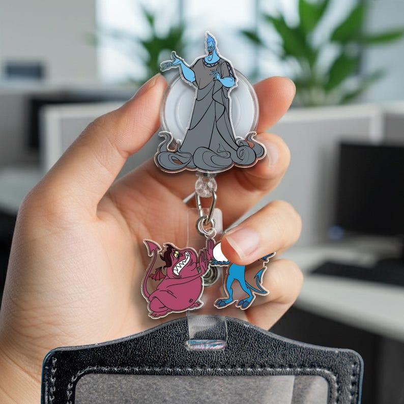 May include: A retractable badge reel featuring cartoon characters. The top character is a blue figure with grey robes. Below are two smaller cartoon figures, one pink and one blue. The reel is attached to a black badge holder.